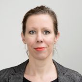 Porträt von Mag. Katharina Katzer mit hochgesteckten Haaren und auffälligem Lippenstift vor hellem Hintergrund.