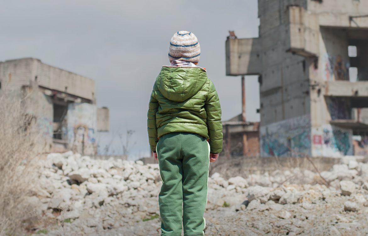 Das Bild zeigt ein Kind, das in Winterkleidung mit dem Rücken zur Kamera vor zerstörten Häusern steht.