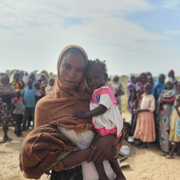 RS118275_20241005_143742 (1) Eine Frau in einem braunen Kopftuch hält ein Kleinkind in bunter Kleidung, umgeben von Gemeinschaftsmitgliedern in einer von Klimawandel und Konflikt betroffenen Region Afrikas.