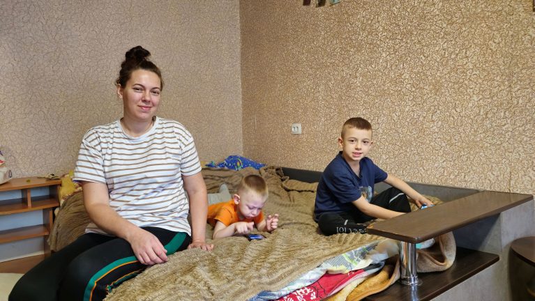 Familie in temporärer Unterkunft in der Ukraine. Mutter in gestreiftem Shirt und zwei Kinder auf einer Couch mit mobilem Tisch