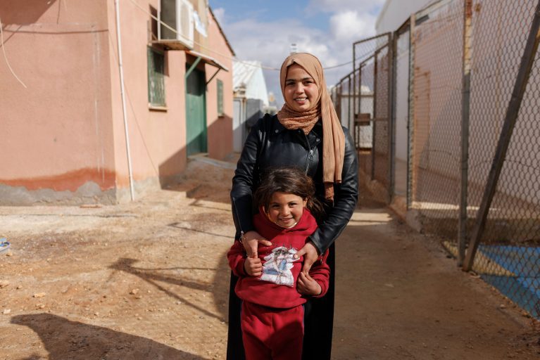 Mutter und Kind in einem Flüchtlingslager in Jordanien. Die Mutter trägt Hijab und Lederjacke, das Kind einen roten Pullover.