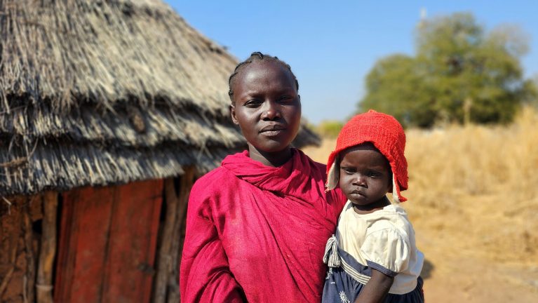 Hütte im Südsudan, davor eine Frau in leuchtend rotem Tuch mit einem Kleinkind.