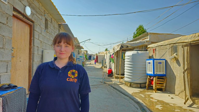 CARE-Mitarbeiterin in blauem Poloshirt steht in einer provisorischen Siedlung mit einfachen Betonbauten und Zelten unter klarem Himmel.