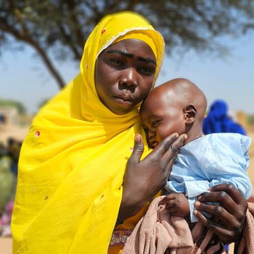 Frau im leuchtend gelben Tuch hält ein Baby schützend im Arm; draußen unter einem Baum, weitere Menschen im unscharfen Hintergrund.