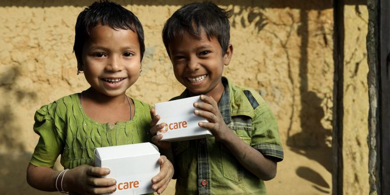 Zwei Kinder halten CARE‑Hilfspakete vor einer Lehmwand – Symbol für schnelle humanitäre Nothilfe durch CARE Österreich.