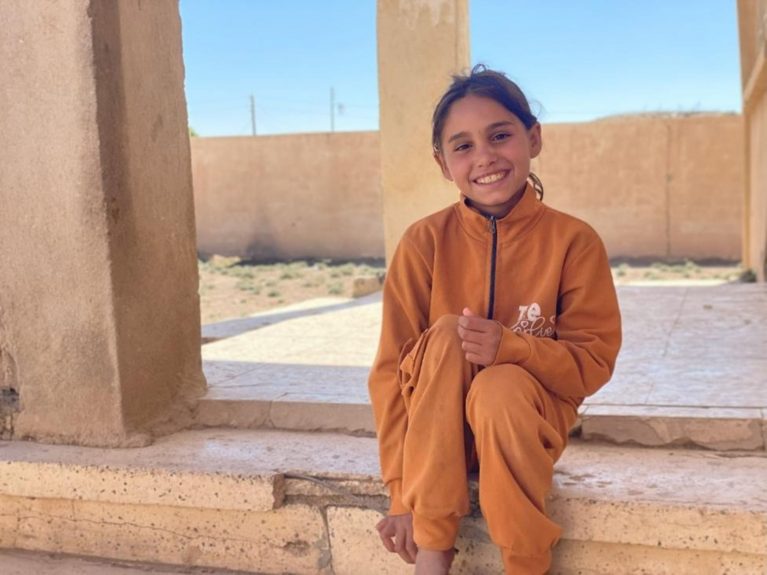 Junges Mädchen in orangefarbener Jacke sitzt lächelnd vor einem Haus in Syrien.