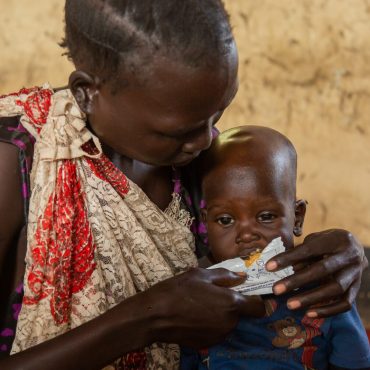 Mutter versorgt ihr Kleinkind in einer einfachen Notunterkunft; im Hintergrund stehen Kartons mit Hilfsgütern. CARE unterstützt Familien im Südsudan, Sudan und Geflüchtete im Tschad mit humanitärer Hilfe.
