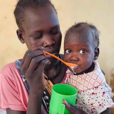 Mutter hält ihr Kleinkind in einer CARE- Gesundheitsstation im Südsudan an der Grenze zum Sudan, während das Kind aus einem grünen Becher trinkt; im Hintergrund steht ein Hygienebehälter mit Wasser.