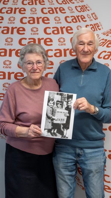 Zwei ältere Geschwister und ehemalige CARE-Paket-Empfänger stehen im CARE Österreich-Büro in Wien vor einer Wand mit CARE-Logo und halten ein historisches Schwarz-Weiß-Foto, auf dem sie als Kinder gemeinsam mit anderen Kindern ein CARE-Paket aus den USA tragen.