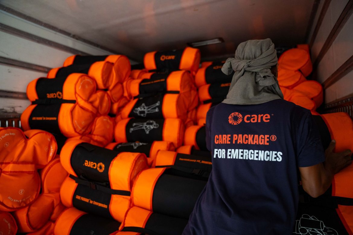 CARE-Helfer vor gestapelten orange-schwarzen Nothilfe-Paketen in einem Transportfahrzeug für Katastropheneinsätze.