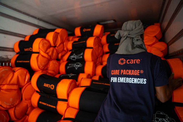 CARE-Helfer vor gestapelten orange-schwarzen Nothilfe-Paketen in einem Transportfahrzeug für Katastropheneinsätze.