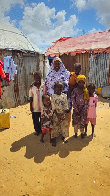 Afrikanische Mutter mit Kopftuch und fünf lächelnden Kindern in farbenfroher Kleidung (orange, rosa, violett) vor Wellblechhütten im Flüchtlingslager.