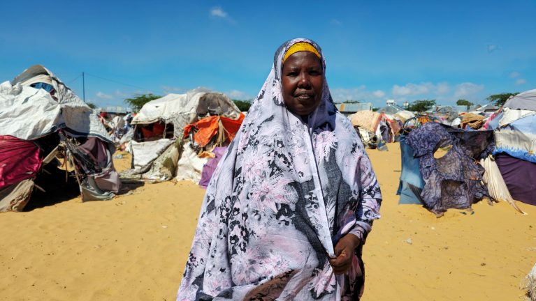 Afrikanische Frau mit violettem Kopftuch und gelbem Stirnband steht frontal vor großem Flüchtlingslager mit hunderten bunten Zelten und Provisorien auf sandiger Ebene