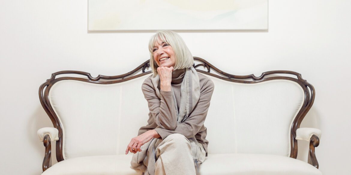 Erika Pluhar, Kunstschaffende und CARE-Testamentsspenderin, sitzt lächelnd auf einem hellen Sofa und stützt ihr Kinn auf die Hand.