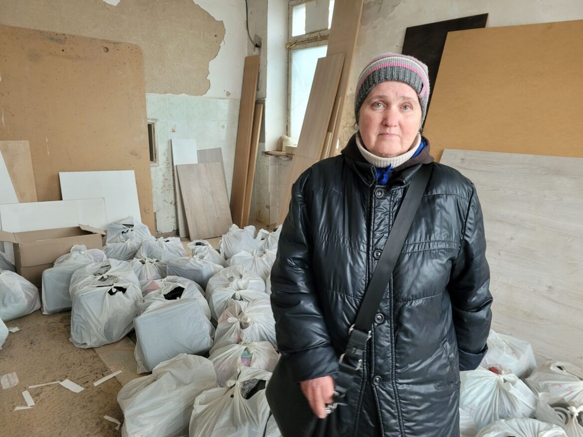 Frau in schwarzer Winterjacke steht in einem unrenovierten Raum vor gestapelten weißen Säcken und Holzplatten